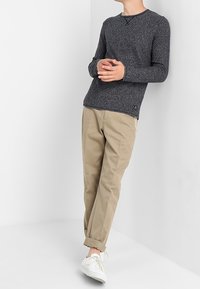 Pull à manches longues gris foncé avec un motif texturé, col rond et emblème latéral, assorti à un pantalon beige et des baskets blanches.