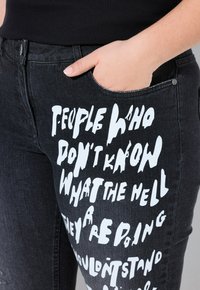 Los jeans de mezclilla negra presentan un gran texto gráfico blanco impreso en la parte frontal. La tela tiene una textura visible y sutiles efectos de decoloración.