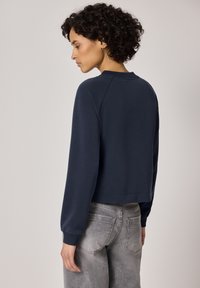 Femme aux cheveux bouclés portant un chemisier bleu marine à manches longues et un jean gris, vue de profil sur un fond neutre.