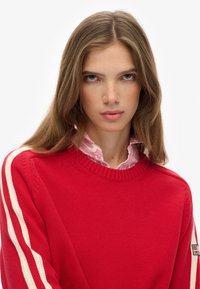 Jonge vrouw met lang bruin haar in een rode trui met witte strepen op de mouwen en een roze gestreept hemd met kraag eronder.