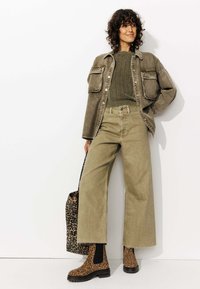 Femme portant un pull vert olive, un jean large verte délavé, une veste marron surdimensionnée, des bottes imprimé léopard, et tenant un sac assorti en imprimé léopard.