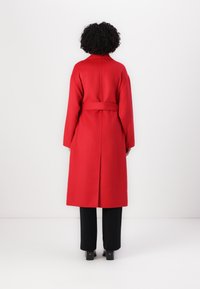 Roter Wollmantel mit tailliertem Gürtel, langen Ärmeln und geradem Schnitt. Verfügt über einen Rückenbelüftungsschlitz und ein klassisches Kragen-Design. Kombiniert mit schwarzen Hosen.