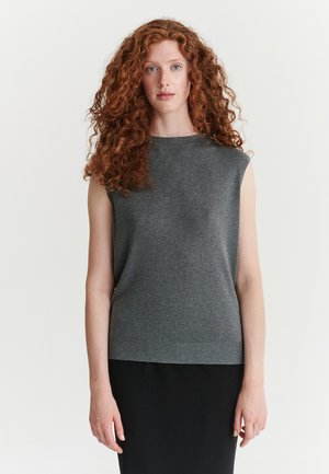 Femme aux longs cheveux roux bouclés, portant un haut gris sans manches et une jupe noire, debout devant un fond blanc.