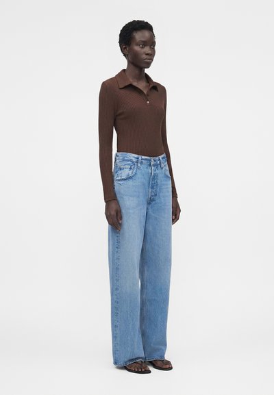Γυναίκα που φοράει καφέ ριπ μπλούζα με μακριά μανίκια και γιακά και μπλε τζιν με ψηλή μέση και φαρδιά πόδια, στέκεται με τα χέρια χαλαρά δίπλα στο σώμα.