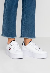 Baskets blanches à plateforme avec une texture en cuir lisse, dotées d'un bout arrondi, d'un laçage à l'avant et d'accents bleu marine et rouge sur le talon.