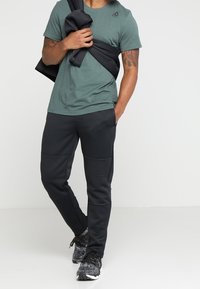 Grön t-shirt med korta ärmar, svarta träningsbyxor med texturerade paneler och svarta sneakers. Personen står med armarna i kors och håller en jacka.