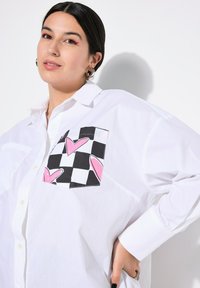 Camicia bianca oversize con bottoni, caratterizzata da una toppa a scacchi bianco e nero con cuori rosa. Tessuto texture, maniche lunghe, design del colletto.