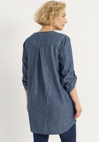 Denim-Tunika-Shirt in Dunkelblau, mit einem Rückenfalten, langen Ärmeln und hochgeschlagenen Manschetten. Glatte Textur mit einem lässigen Design.