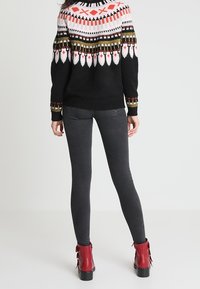 Czarny sweter robiony na drutach z kolorowymi geometrycznymi wzorami, w połączeniu z szarymi legginsami i czerwonymi botkami na obcasie z frędzlami.