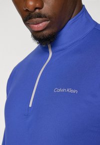 Karališkai mėlyna pusiau užtraukiama palaidinė su šviesiai pilku užtrauktuku ir balta siuvinėta "Calvin Klein" logotipo detalė ant krūtinės. Lygi, tampri medžiaga.