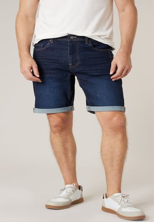 Homme portant un short en jean bleu foncé retroussé, un t-shirt blanc et des baskets blanches à semelles beige, debout sur un fond neutre.
