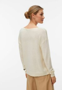 Femme aux cheveux blonds attachés en chignon, portant un pull en maille crème ample et un pantalon beige, vue de dos et légèrement tournée vers la droite.