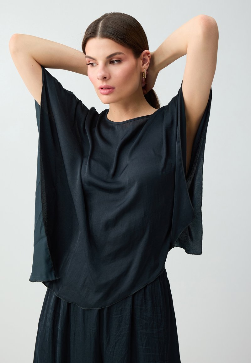 Femme aux cheveux foncés attachés, portant un haut noir ample et une jupe, levant les bras avec les mains derrière la tête, regardant légèrement sur le côté.