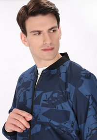 Marine bomberjack met abstracte geometrische patronen in verschillende tinten blauw en zwart. Voorzien van een zwarte kraag en een rits aan de voorkant.