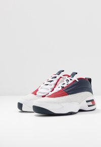 Białe i granatowe skórzane sneakersy z czerwonymi akcentami, teksturowanymi panelami, okrągłym przodem i grubą gumową podeszwą. Zawierają detale z logo oraz sznurowane zapięcie.