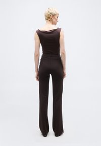 Άτομο με κοντά ξανθά μαλλιά που φοράει αμάνικο σκούρο καφέ τοπ και ψηλόμεσα σκούρα καφέ παντελόνια, στέκεται με το σώμα στραμμένο προς τα πίσω σε λευκό φόντο.