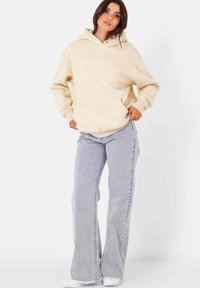Felpa color crema con tasca frontale, abbinata a jeans chiari a campana. La texture è morbida e i jeans hanno orli sfilacciati.