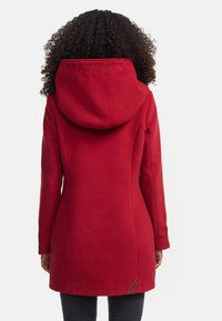 Manteau rouge avec une grande capuche, présentant une broderie de marque. Tissu doux, coupe ajustée et un détail latéral près de l'ourlet.