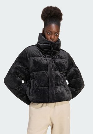 Vrouw met natuurlijk haar draagt een zwarte doorgestikte pufferjas en beige broek tegen een effen lichte achtergrond.