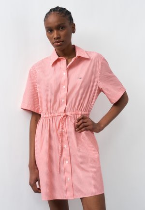 Mujer con vestido de rayas verticales rojas y blancas de manga corta, con botones y cintura con cordón, posando con una mano en la cadera.
