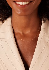 Gouden kettinghalsketting gedragen tegen een huidkleurige achtergrond, aangevuld met een beige pinstriped blazer. De ketting heeft een delicate, minimale ontwerp.