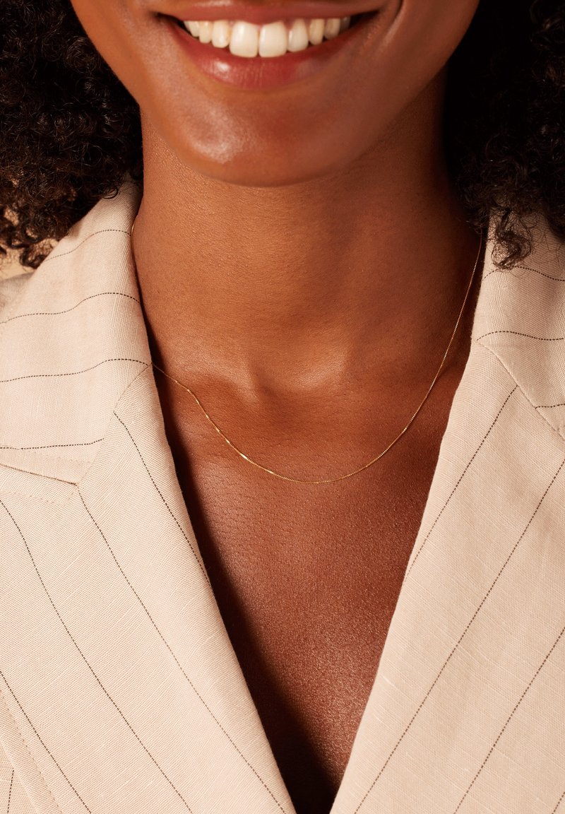 Gouden kettinghalsketting gedragen tegen een huidkleurige achtergrond, aangevuld met een beige pinstriped blazer. De ketting heeft een delicate, minimale ontwerp.