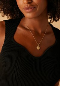 Zwarte geribde tanktop met een ronde halslijn, voorzien van een delicate gouden ketting met een ronde hanger en een textuur aan de oppervlakte.