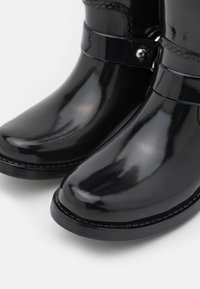 Bottines noires brillantes avec bouts arrondis, lanières décoratives et petits talons sur fond blanc.
