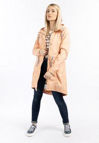 Jachetă impermeabilă de culoare piersică, cu glugă, talie strânsă, accente albastre și închidere cu capse, purtată cu blugi închiși și adidași gri.