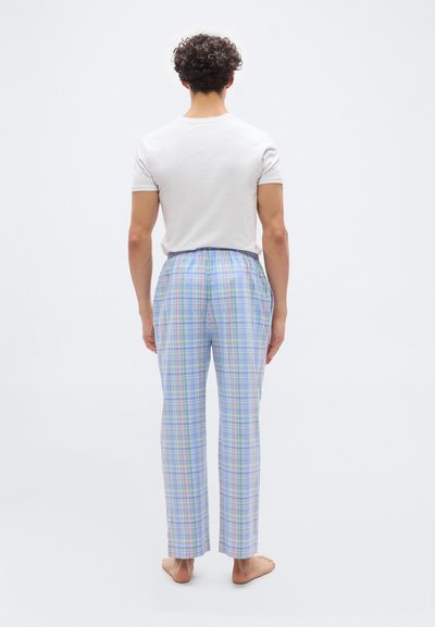 Άνδρας που στέκεται ξυπόλητος, με την πλάτη γυρισμένη, φορώντας λευκό μπλουζάκι και παστό μπλε καρό παντελόνι, σε λευκό απλό φόντο.