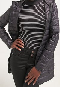 Musta, toppauksella varustettu takki, jossa on hupullinen kaulus, vetoketjusuljenta edessä ja pystysaumat. Päällä mustavalkoraidallinen pooloneule ja korkeavyötäröiset farkut.