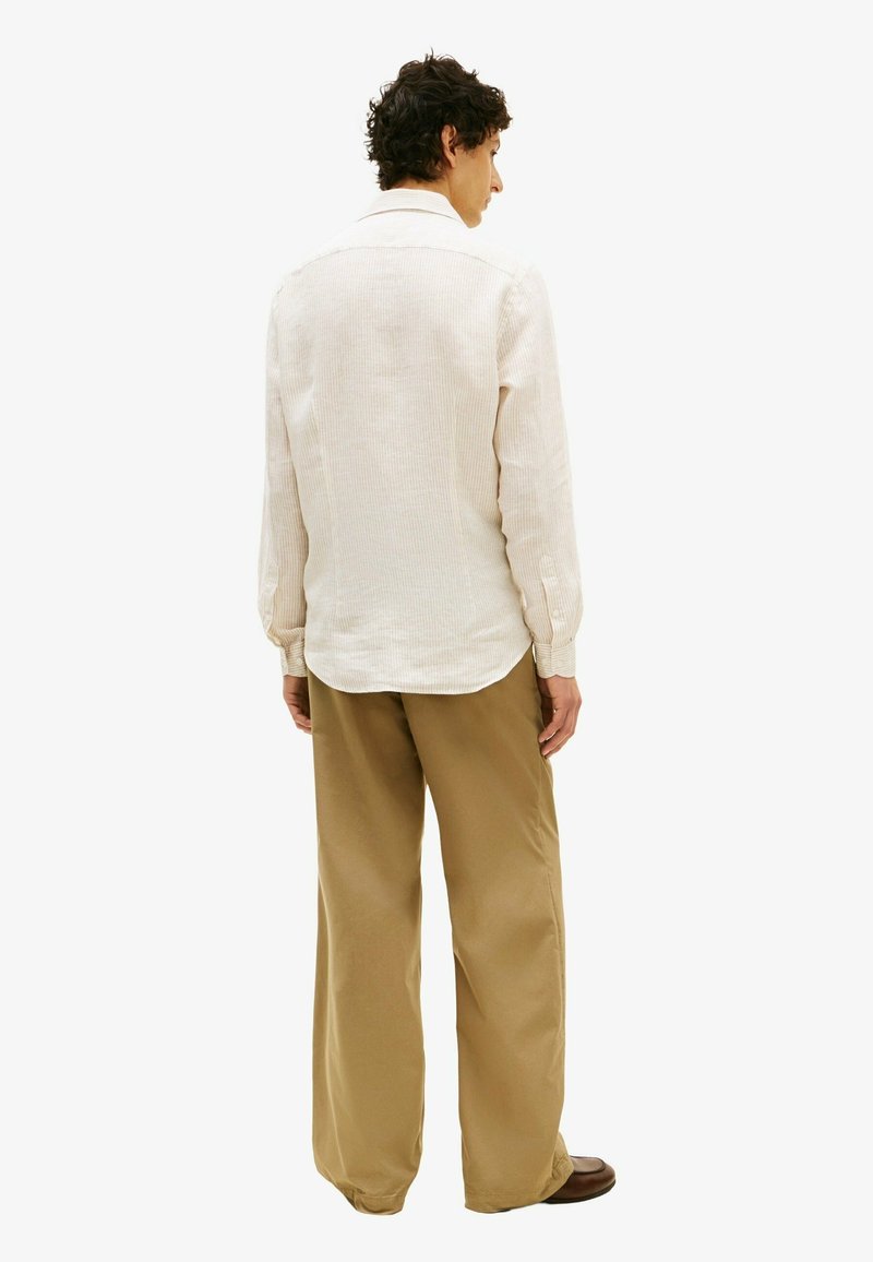 Homme aux cheveux bouclés portant une chemise beige à manches longues, un pantalon large olive et des chaussures marron, debout, tourné de dos sur un fond blanc.