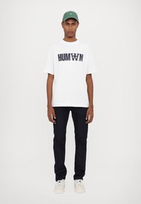 White cotton t-shirt with "HUMAN" text in navy, paired with dark denim jeans and white sneakers. Also wearing a green cap.