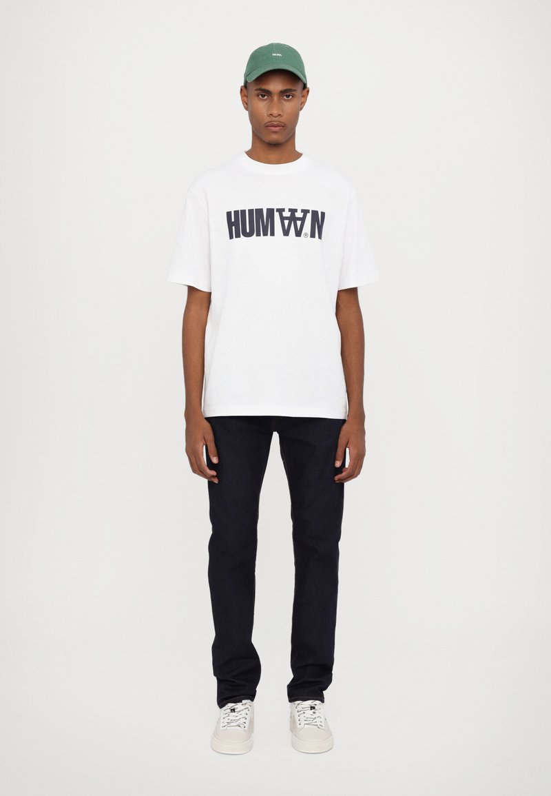 White cotton t-shirt with "HUMAN" text in navy, paired with dark denim jeans and white sneakers. Also wearing a green cap.
