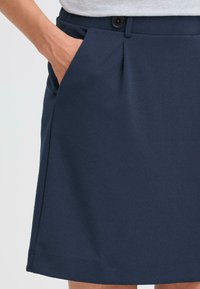 Gonna plissettata blu navy realizzata in tessuto morbido, con un bottone visibile e tasche laterali. L'orlo è rifinito in modo ordinato.