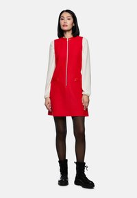 Rotes Kleid mit einem Reißverschluss vorne, ausgestattet mit langen weißen Ärmeln und verzierten Taschen. Getragen mit schwarzen Schnürstiefeln und transparenten schwarzen Strumpfhosen.