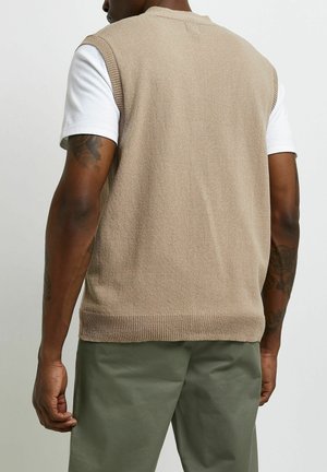 Béžový pletený svetřík bez rukávů s kulatým výstřihem, nošený přes bílou krátkou košili, doplněný olivově zelenými kalhotami.