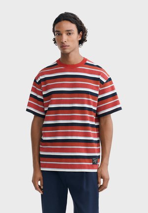 Jeune homme aux cheveux bouclés et foncés, portant un t-shirt rouge, blanc et bleu marine à rayures horizontales et un pantalon sombre, debout devant un fond uni.