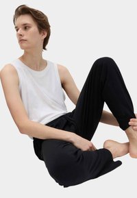 Persona joven con una camiseta blanca sin mangas y pantalones negros, sentada descalza con una pierna levantada y mirando hacia un lado sobre un fondo liso.