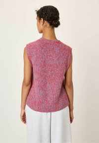 Žena s vrkočom vo vlase, oblečená v bezrukávovom pletenom svetríku v červenej a fialovej farbe a svetlosivých nohaviciach, zobrazená zozadu.