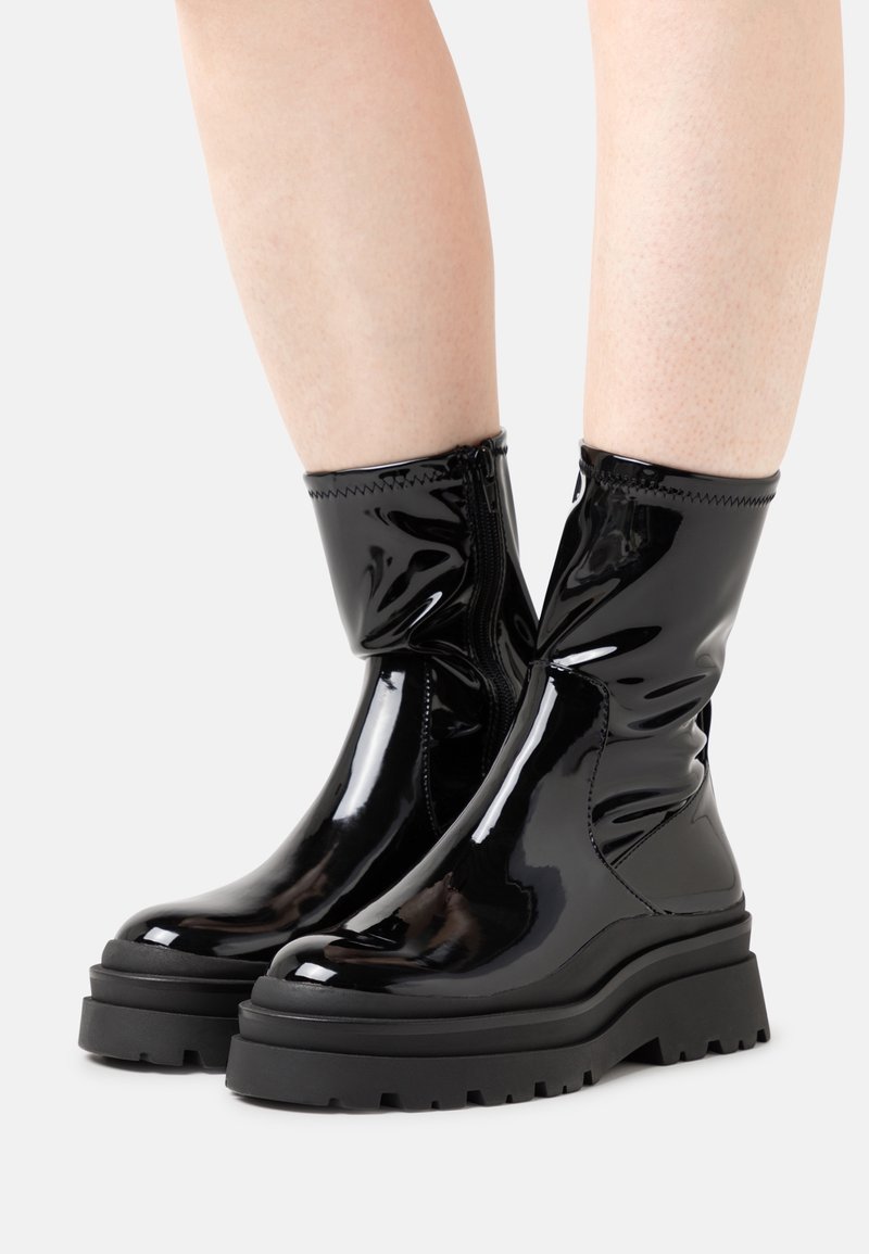 Bottes noires brillantes mi-mollet avec semelles à relief massif portées sur jambes nues sur fond blanc.