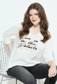 Donna con lunghi capelli castani che indossa una maglietta bianca a maniche lunghe con testo e jeans scuri, seduta su una sedia di metallo a rete contro uno sfondo bianco.