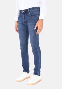 Uomo che indossa jeans blu attillati, camicia bianca e sneakers bianche, in piedi davanti a uno sfondo chiaro e uniforme.