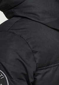 Chaqueta puffer negra con una textura suave, que presenta un parche de logo redondo en la manga y costuras visibles a lo largo de las uniones.