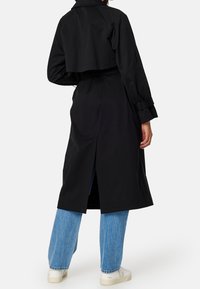 Černý trenčkot s krátkým střihem, dlouhými rukávy a rozparkem na zádech. Nošený přes modré džíny s volnějším střihem a bílé tenisky.