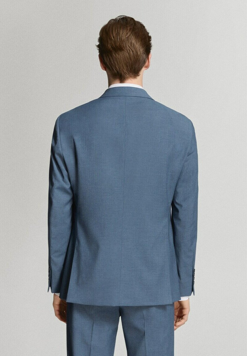 Hombre con traje azul a medida de espaldas frente a una pared lisa de color gris claro, mostrando la parte trasera de la chaqueta y los pantalones.