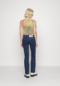 En blond person som bär en genomskinlig blommig topp, mörkblå jeans med "RAGGED"-etikett och vita stövlar, står vänd bort från kameran mot en enkel bakgrund.