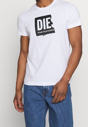 Mand iført hvid kortærmet T-shirt med sort-hvidt mærke med teksten "DIES FOR SUCCESS" og blå jeans mod ensfarvet baggrund.