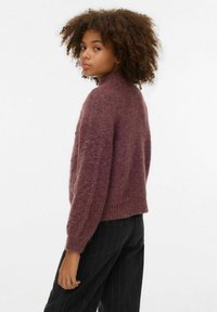 Apaļš kakls, pūkains tumši sarkans džemperis ar ribotu apakšu un garām piedurknēm, kombinēts ar augsta vidukļa melnām svītrainām biksēm, redzams tekstūras detaļas.