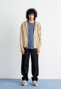 Sweatshirt bege com fecho, camiseta azul, jeans pretos de perna larga, sapatilhas cinzentas. Roupa casual com bolsos e detalhes em costura contrastante.